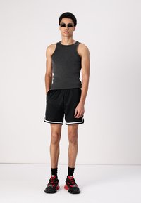 Shorts de sport noirs avec des accents blancs, débardeur gris, lunettes de soleil noires et baskets noires épaisses avec des détails rouges. Fond uni.