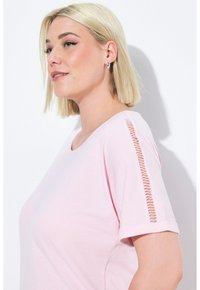 Světle růžové tričko vyrobené z měkké látky, které má kulatý výstřih a výrazné boční rozparky. Otočené rukávy dodávají detail a design.