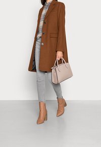 Borsa a mano beige, cappotto marrone con colletto a scollo, camicia a quadri chiari, jeans skinny grigi e stivali alla caviglia con tacco color sabbia. Texture lisce, vestibilità su misura.