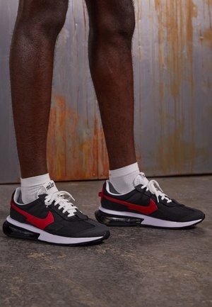 Ben iført sorte og røde Nike-sneakers med hvide snørebånd og hvide sokker, stående på en struktureret betongulv foran en rustet metalvæg.
