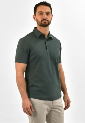 Polo-Shirt in dunklem Grün, kurzärmlig, glattes Material, Knopfleiste mit drei Knöpfen, dezentes Logo-Detail auf der Brust, kombiniert mit hellen Hosen.