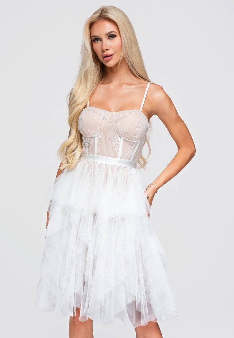 Femme blonde portant une robe corset en dentelle blanche avec jupe en tulle superposée, posant les mains dans les poches sur un fond uni.
