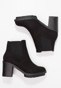 Ein Paar schwarze Wildlederstiefeletten mit klobigen Absätzen und elastischen seitlichen Einsätzen auf einem weißen Hintergrund.