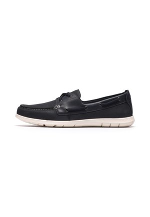 Chaussure bateau décontractée en cuir noir avec semelle en caoutchouc blanche et design sans lacets, vue de côté sur fond blanc.