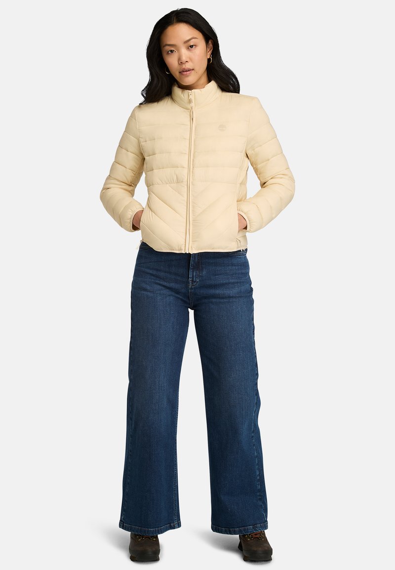 Giacca a puffo beige chiaro con colletto alto, caratterizzata da cuciture orizzontali e a chevron, abbinata a jeans blu a gamba larga e stivali scuri.