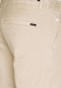 Beige Baumwollhosen mit glatter Textur, einer Gesäßtasche mit schwarzem Etikett und weißen Nähten.