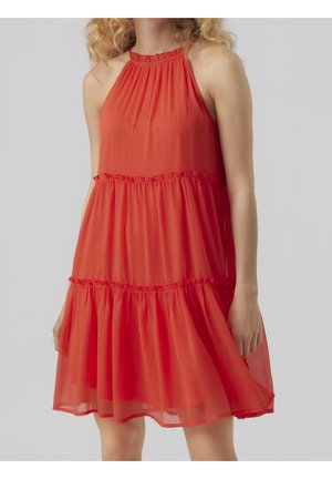 Femme portant une robe rouge sans manches, longueur genou, à volants superposés froncés et col montant, sur fond gris uni.