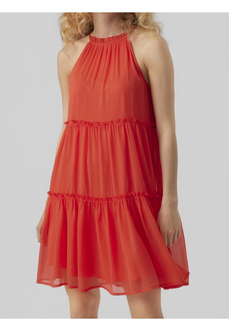 Femme portant une robe rouge sans manches, longueur genou, à volants superposés froncés et col montant, sur fond gris uni.