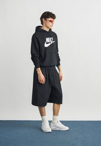 Νεαρός άνδρας με μαύρο φούτερ Nike, μαύρα φαρδιά σορτς, λευκές κάλτσες, λευκά αθλητικά παπούτσια και γυαλιά ηλίου με κόκκινους φακούς, στέκεται σε μπλε χαλί.