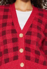 Punainen ja tumma viininpunainen ruudullinen neulecardigan, jossa on V-pääntie, teksturoitu neule ja viisi suurta vaaleaa nappia edessä.