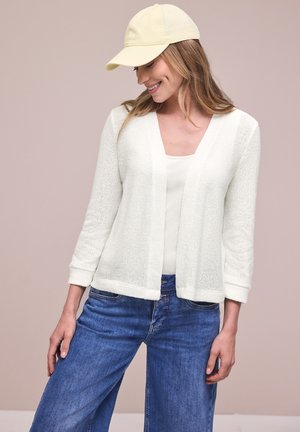 Kobieta ubrana w biały dziergany kardigan, biały top, niebieskie dżinsy i bladożółtą czapkę z daszkiem, uśmiechająca się i patrząca w dół na lewo.