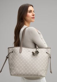 Borsa a secchiello beige con motivo in rilievo, accenti in pelle marrone, doppi manici e portachiavi a forma di fiore decorativo. Finitura testurizzata.