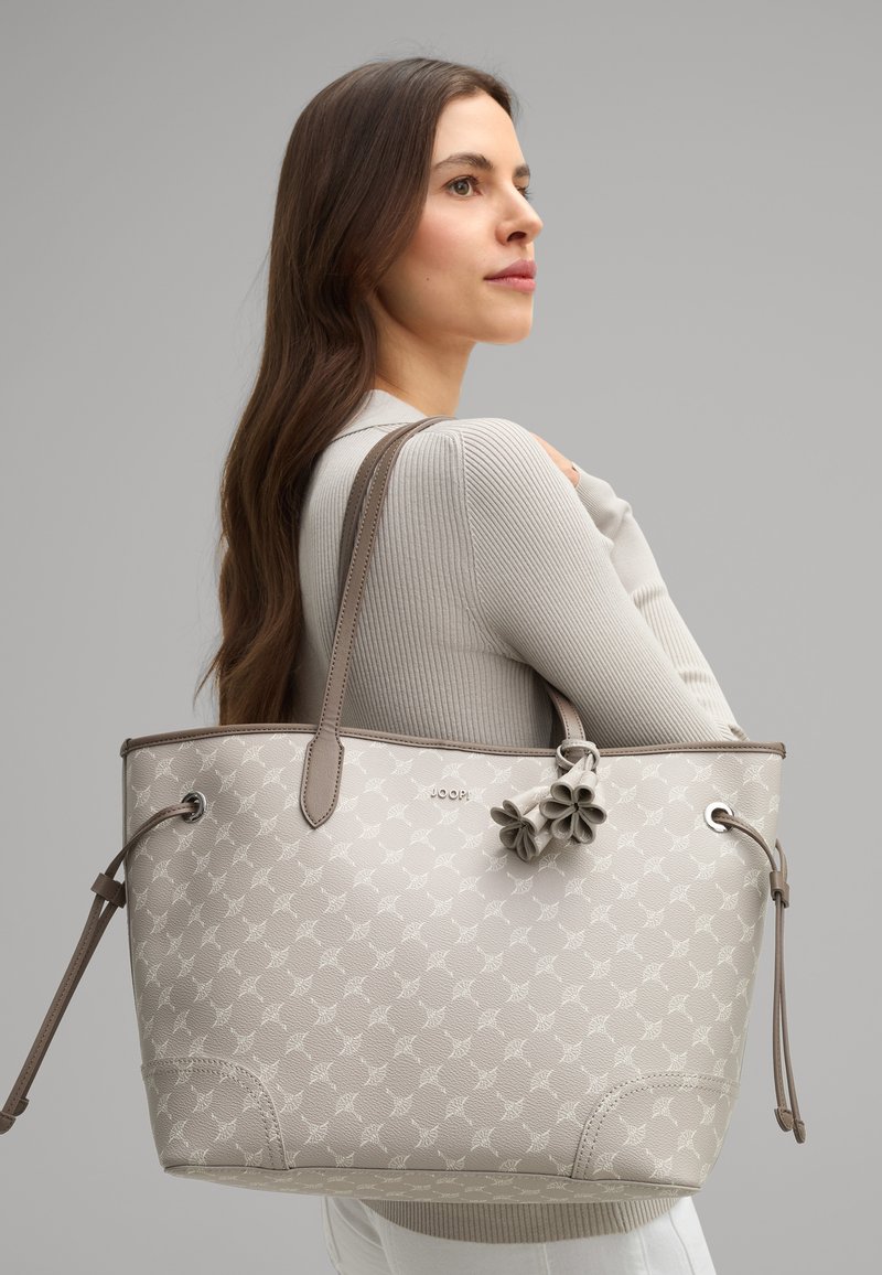 Borsa a secchiello beige con motivo in rilievo, accenti in pelle marrone, doppi manici e portachiavi a forma di fiore decorativo. Finitura testurizzata.