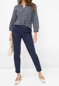 Bluzka w granatowy wzór w okrągłe kropki, z zapięciem na guziki z przodu i rękawami 3/4. Slimowane granatowe spodnie i jasnobeżowe baleriny, trzymająca torebkę w kolorze kremowym.
