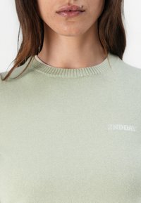 Hele roheline kootud pullover, millel on ümmargune kaelus, soonikservad ja väike valge logo vasakul rinnapiiril. Pehme tekstuur.