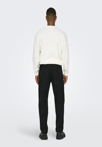 Maglione bianco lavorato a maglia con scollo rotondo, abbinato a pantaloni cargo neri con tasche laterali e una texture liscia. Scarpe nere completano il look.