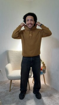 Hombre sonriente con cabello rizado ajustándose la sudadera marrón, de pie sobre un suelo alfombrado cerca de un sillón blanco y una mesa pequeña con un jarrón y una taza.