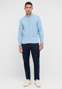Jasnoniebieska koszula zapinana na guziki z kołnierzykiem i długimi rękawami, w zestawieniu z ciemnoniebieskimi dżinsami i beżowymi trampkami. Gładka, jednolita tkanina.