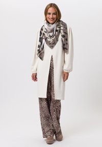 Witte jurk met lange mouwen en een split aan de voorkant, gecombineerd met beige broek met luipaardpatroon en een doorzichtige sjaal met een grijs en zwart dierenprint.