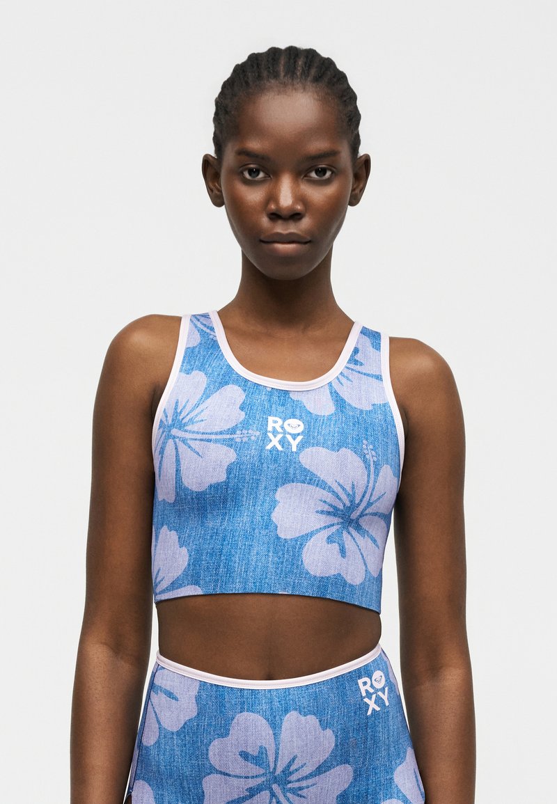 Femme aux cheveux tressés portant un haut court bleu à fleurs et un bas taille haute assorti avec une bordure blanche et le logo ROXY.
