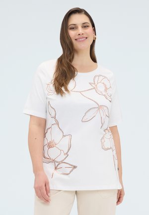 Frau mit langen braunen Haaren, die ein weißes, kurzärmliges Oberteil mit großem orangefarbenem Blumen-Umrissmuster und beige Hose trägt, lächelt vor hellem Hintergrund.