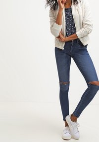 Krämfärgad bomberjacka över en marinblå t-shirt med fågelmönster, kombinerad med medelhöga, smala mörkblå jeans med knäskärningar och vita sneakers.