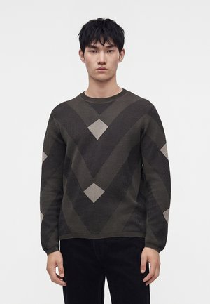 Giovane uomo con capelli neri corti che indossa un maglione scuro con fantasia e pantaloni neri, in piedi davanti a uno sfondo chiaro e semplice.
