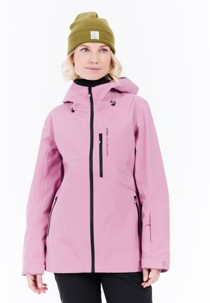 Vrouw in een roze gewatteerde jas met zwarte ritsen en een olijfgroen mutsje, staande tegen een witte achtergrond.
