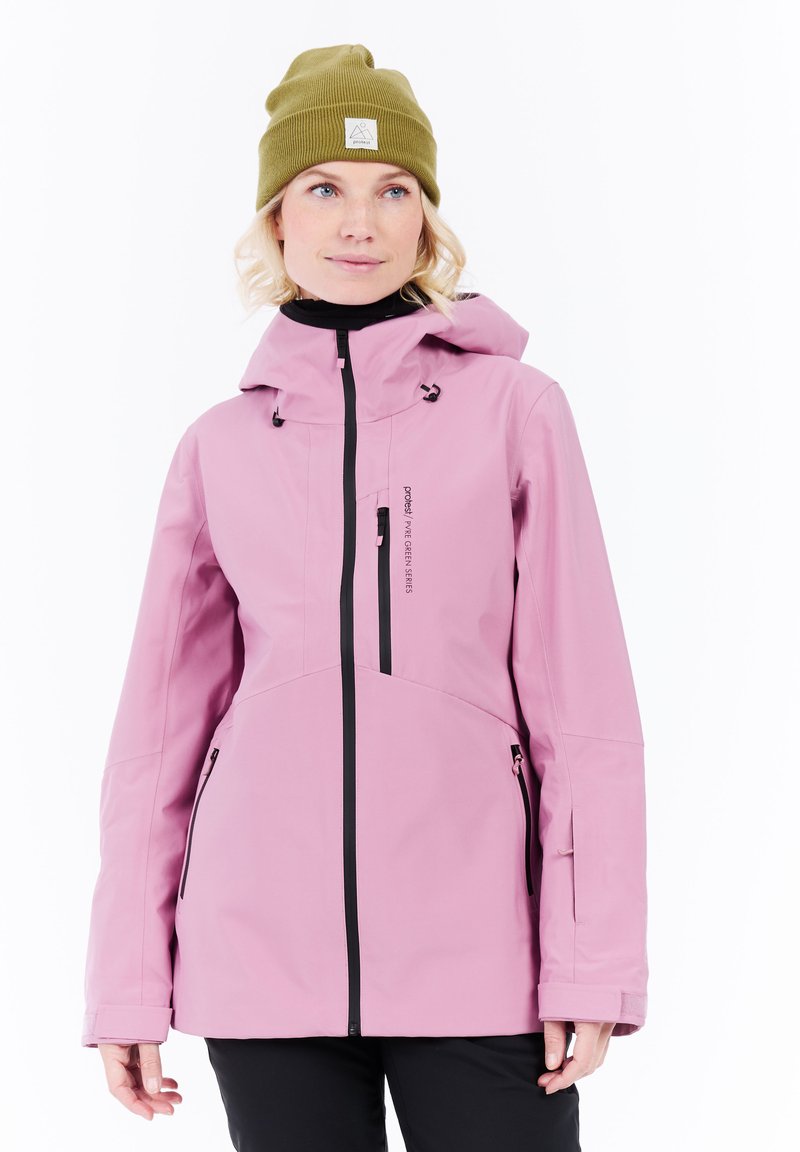 Woman wearing a pink hooded jacket with black zippers and an olive green beanie, standing against a white background.