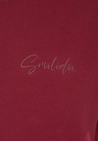 Tessuto bordeaux con un motivo lavorato a maglia testurizzato, che presenta il nome del marchio "Smilodox" in carattere corsivo grigio chiaro al centro.