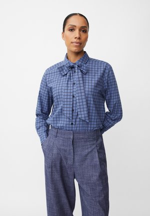 Blusa a quadri blu con colletto a fiocco e bottoni neri, abbinata a pantaloni texture blu scuro con pieghe frontali.