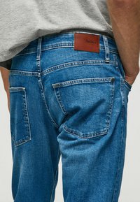 Mėlyni džinsai su oda aptrauktu užpakaliniu juosmens kraštu, kuriame yra "Pepe Jeans" logotipas, taip pat matomomis siūlėmis ant kišenių.