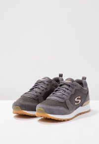 Szare buty sportowe z siateczkowymi i zamszowymi materiałami, z białą podeszwą pośrednią, gumową podeszwą i subtelnym logo po boku.