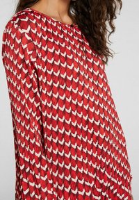 Robe plissée rouge dotée d'un motif géométrique blanc. Les manches longues et le col rond ajoutent à son design fluide.