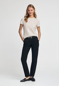 Witte katoenen t-shirt met korte mouwen, met een klein logo. Gecombineerd met zwarte broek en zwarte instappers, staand tegen een lichte achtergrond.