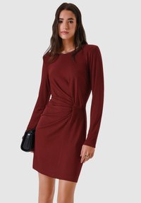 Abito bordeaux a maniche lunghe con dettaglio nodo intrecciato in vita. Texture liscia, collo tondo e silhouette aderente. Accessorio borsa nera.