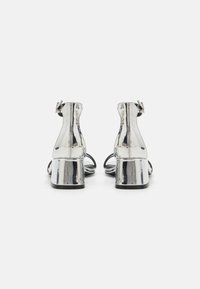 Sandali metallici argentati con tacco e cinturini alla caviglia, caratterizzati da una superficie liscia, tacchi a blocco e un design minimalista. La vista posteriore mette in evidenza i dettagli della cucitura.
