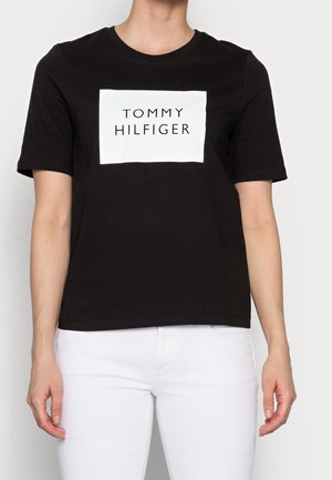 Camiseta de algodón negra con un parche rectangular blanco que lleva el texto en negro "TOMMY HILFIGER". Cuello redondo y manga corta, corte clásico.
