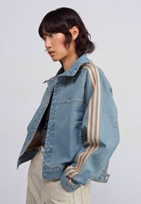 Gaiši zilas džinsa jaka ar lielu piegriezumu, ar divām vertikālām beidža krāsas svītrām gar piedurknēm un klasisku apkakles dizainu.