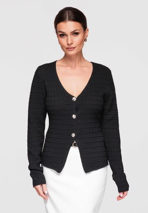 Femme portant un cardigan noir texturé à quatre boutons et une jupe blanche, posant devant un fond gris clair uni.