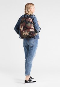 Mochila camuflada con un exterior de tela, que presenta tonos de marrón y verde. Tiene un bolsillo frontal y correas ajustables. Se lleva sobre unos vaqueros.