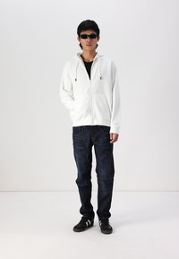Haut à zip blanc avec cordons, associé à un jean en denim foncé et des baskets noires. Le modèle porte des lunettes de soleil noires. Fond uni.