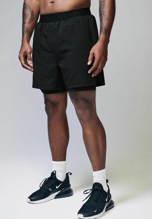 Pantalones cortos deportivos negros con cintura elástica, con un corte casual y bolsillos laterales. Llevados con zapatillas negras y calcetines blancos hasta el tobillo.