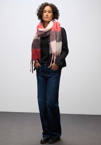 Flerfarvet plaid-scarf med pink, rød og grå nuancer, båret over en sort top, parret med mørkeblå jeans, stående på et gråt gulv.
