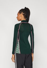 Mørkegrønn langermet topp med høy krage, rosa trim langs sømmene, og et mønstret sidepanel i lys grønn og mørkegrønn.