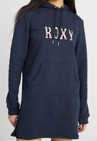 Mörkblå hoodieklänning med känguruficka och dekorativ "ROXY"-text i kamouflage-mönster. Tillverkad av mjukt material.