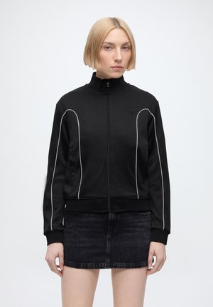 Femme aux cheveux blonds courts portant une veste zippée noire avec des bandes blanches et une jupe courte en jean noire, debout devant un fond blanc.