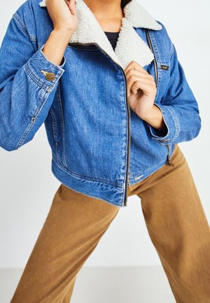 Person wearing blue denim jacket with white sherpa collar and tan pants, adjusting the jacket zipper.
