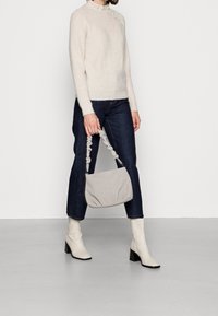 Sac à main gris avec un design froncé et une lanière texturée. Le modèle porte un pull en crème côtelé et un jean foncé, associé à des bottes blanches à talons.