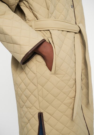 Beige quilted coat featuring a belted waist, contrast brown accents, and side pockets. The fabric has a soft texture with a diamond pattern.